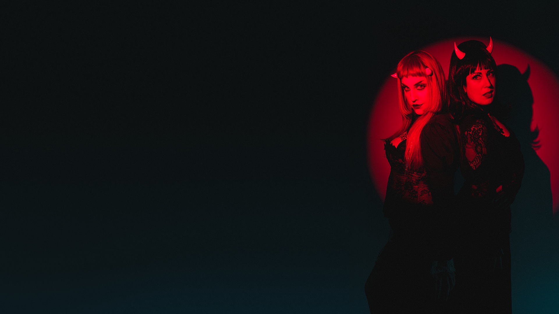 Models Stormy September and Ruby Lamb pose in gothic attire in a red spotlight wearing bone white imp and incubus devil horns. Photo by Matt Stone.