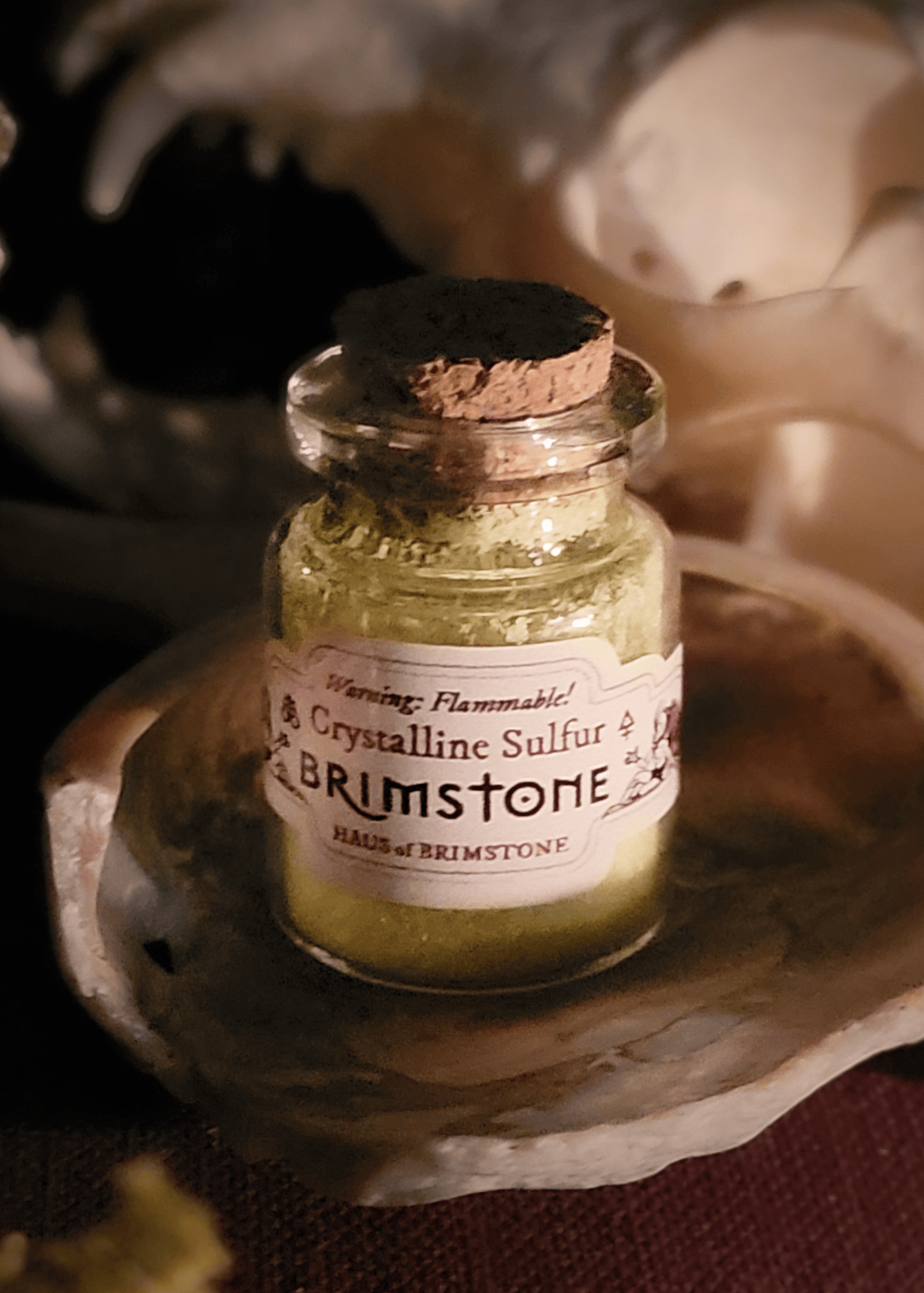 A tiny bottle of crystallized sulfur with a cork stopper sits on a cowry shell with a fox skull visible in the background. The label reads, “Warning: Flammable! Crystalline Sulfur. Brimstone. Haus of Brimstone.” The label also depicts two devils and two different alchemical symbols for brimstone.