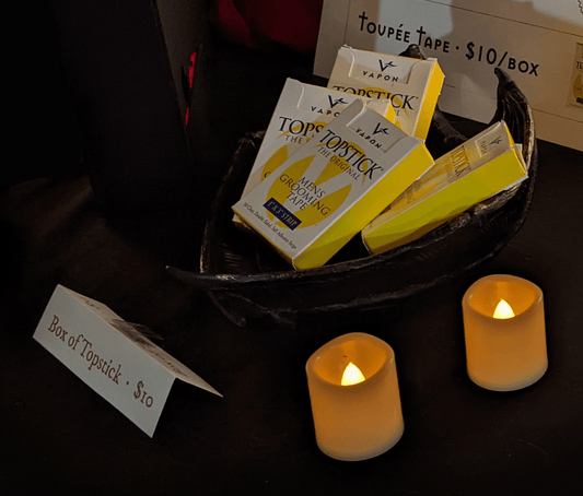 Boxes of Topstick toupee tape sit in a gothic winged bowl next to two candles. Sign reads “Box of Topstick: $10”